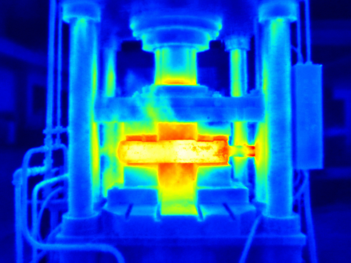 Thermal image of a forging press showing a white-hot steel billet and localised overheating at the right-hand guide bushing.
