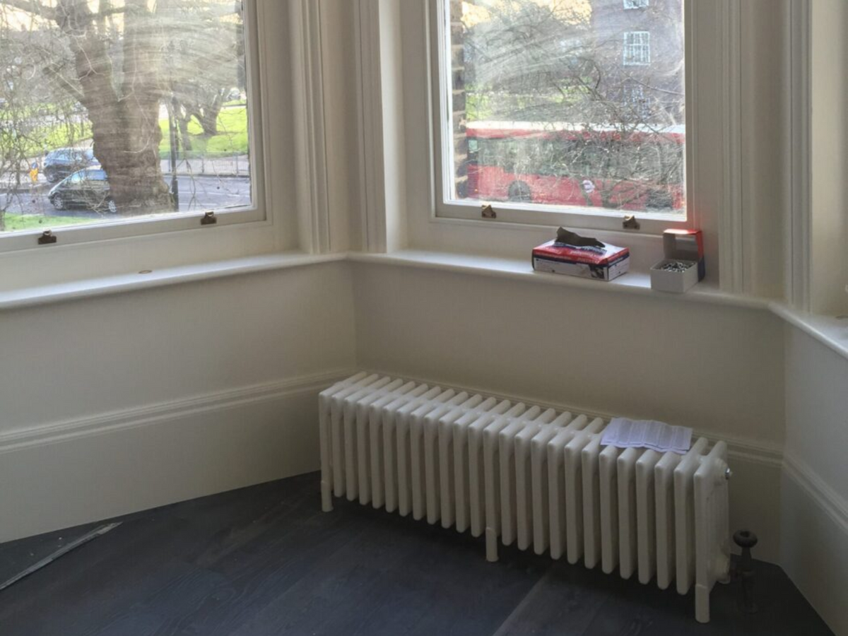 A white vintage-style radiator positioned against an inner corner wall beneath two large windows, with a dark wood floor and a street scene visible outside.